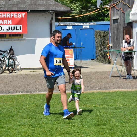 Ein Mann in einem blauen Shirt mit der Nummer 109 läuft mit einem kleinen Mädchen auf einem Rasenfeld. Hinter ihnen sind Fahrräder, ein Gebäude mit einem roten Banner und ein Tisch mit einem Mann.