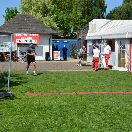 Ein Läufer nimmt an einem Marathon-Event vor einem Gebäude mit dem Schild 'Feuerwehrfest Ossarn' teil. Mehrere Leute stehen herum, und ein Zelt ist in der Nähe aufgestellt.