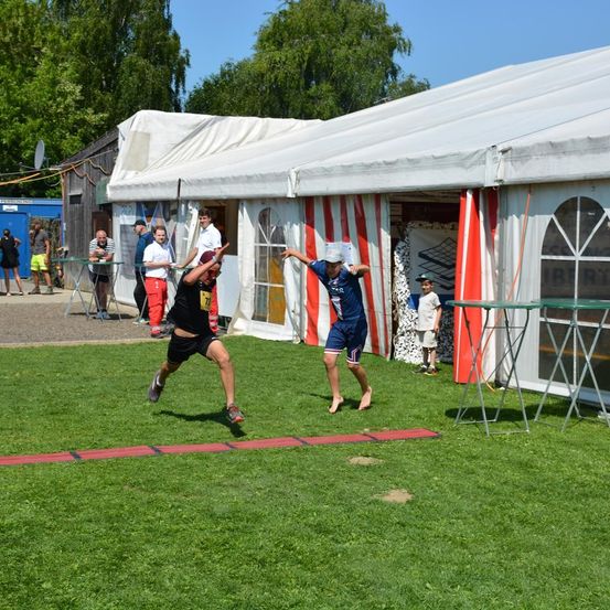 Zwei Athleten nehmen an einem Laufwettbewerb auf einem Rasenfeld teil. Zuschauer versammeln sich in der Nähe eines Zelts mit rot-weiß gestreiften Vorhängen.