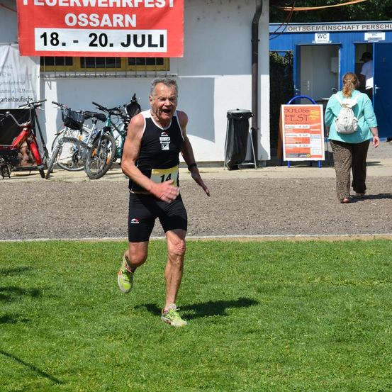 Ein Mann läuft einen Marathon und trägt eine Startnummer 14. Eine Frau mit Rucksack läuft im Hintergrund. Dahinter stehen Fahrräder und ein Gebäude.
