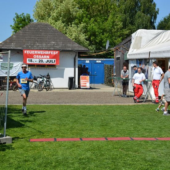 Ein Mann läuft auf einem Rasenfeld, wahrscheinlich an einem Marathon teilnehmend. Hinter ihm stehen mehrere Personen in der Nähe von Zelten und einem Gebäude mit einem Schild, das 'Feuerwehrfest Ossarn 18-20 Juli' besagt. In der Nähe sind Fahrräder geparkt.