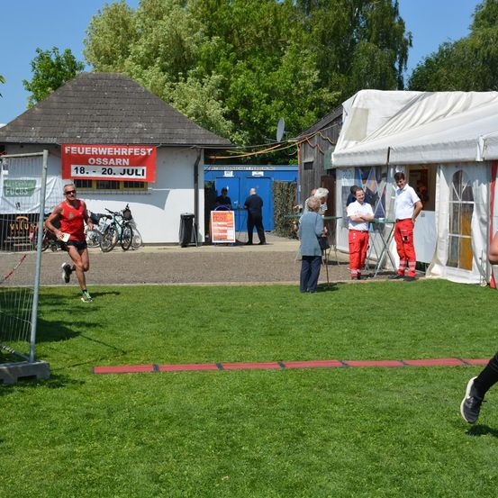 Ein Läufer in einem roten Trikot läuft an einem Gebäude mit einem Schild vorbei, das Feuerwehrfest Ossarn anzeigt. Mehrere Personen stehen vor einem Zelt und einem weiteren Gebäude.