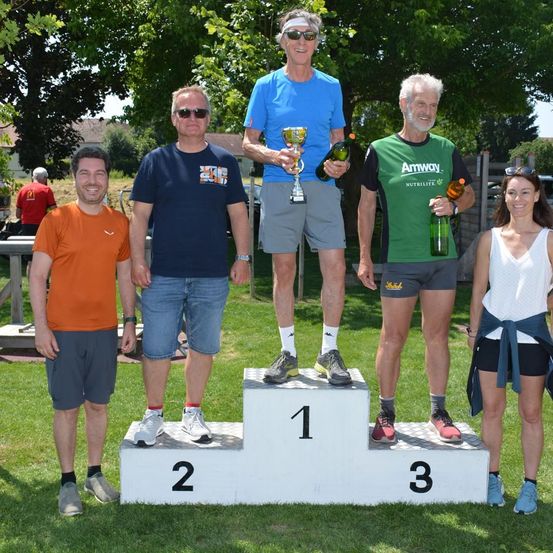 Fünf Personen stehen auf einem Podest mit grünen Flaschen, tragen Turnschuhe und Freizeitkleidung. Eine Person hält eine Trophäe, eine andere eine grüne Flasche, und drei weitere stehen hinter ihnen. Bäume und ein Zaun sind im Hintergrund.