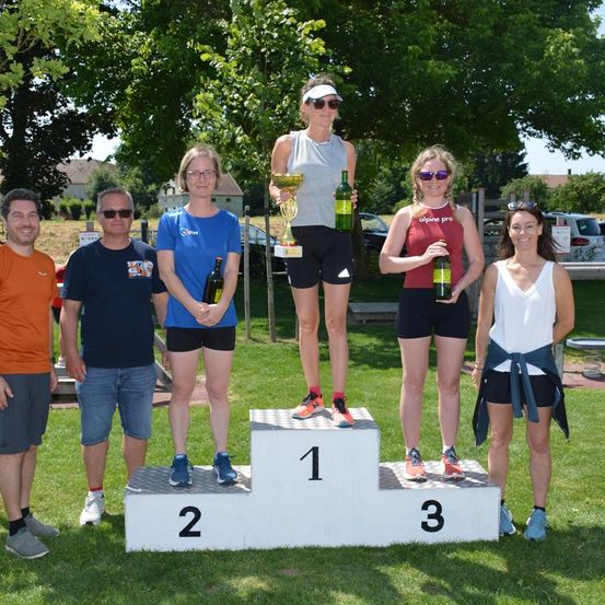 Eine Gruppe von Menschen steht auf einem Podium in einem Park, zwei halten Trophäen und Flaschen. Die Frau in der Mitte trägt eine Medaille und eine Kappe. Hinter ihnen stehen drei Männer links und eine Frau rechts.