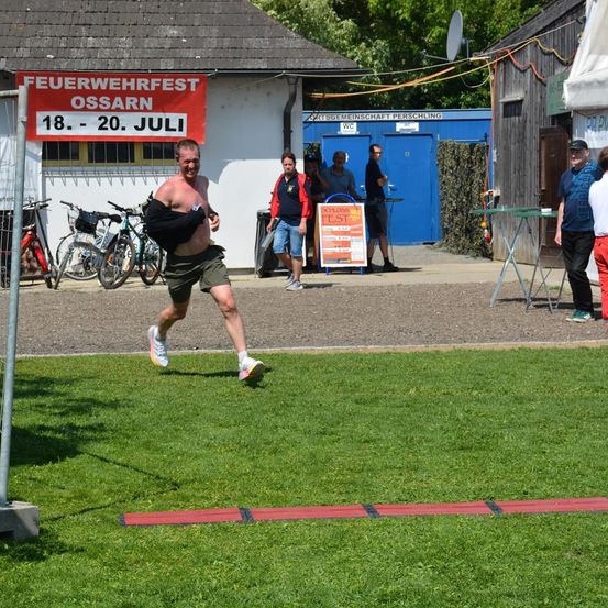 Ein Mann läuft ohne Hemd auf einem Rasenfeld, mit einem roten Banner, das Feuerwehrfest Ossarn besagt. Fahrräder sind geparkt und Leute stehen in der Nähe eines Gebäudes.