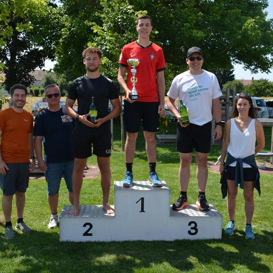 Fünf Personen stehen auf einem Podest unter einem Baum. Die Person in der Mitte hält eine Trophäe. Zwei andere an den Seiten halten Flaschen. Einer trägt ein rotes Shirt und der andere ein weißes Shirt mit Text. Die Person auf der linken Seite trägt ein oranges Shirt.