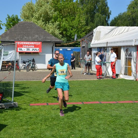 Eine Läuferin mit der Nummer 19 läuft auf einem Rasenfeld mit roten Linien. Hinter ihr folgt ein Mann mit Sonnenbrille. Mehrere Personen stehen in der Nähe eines Zeltes und eines Gebäudes mit der Aufschrift Feuerwehrfest Ossarn.