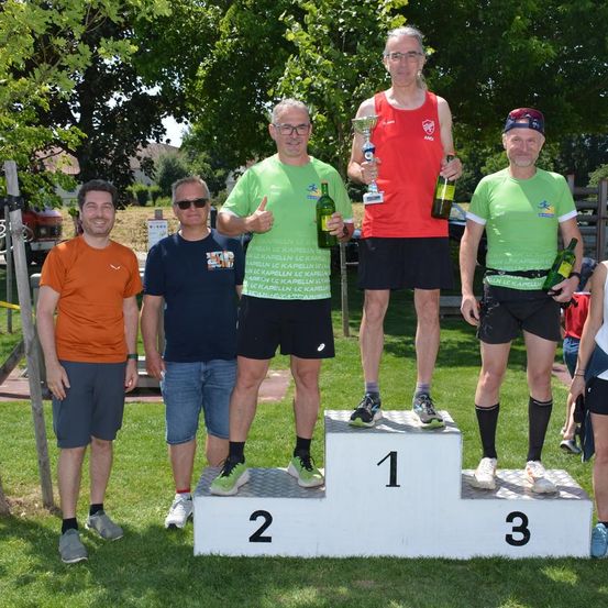Fünf Männer stehen auf einem Podest, einer in Rot hält einen Pokal und zwei in Grün halten Flaschen. Sie stehen auf einem Rasenfeld mit Bäumen im Hintergrund.