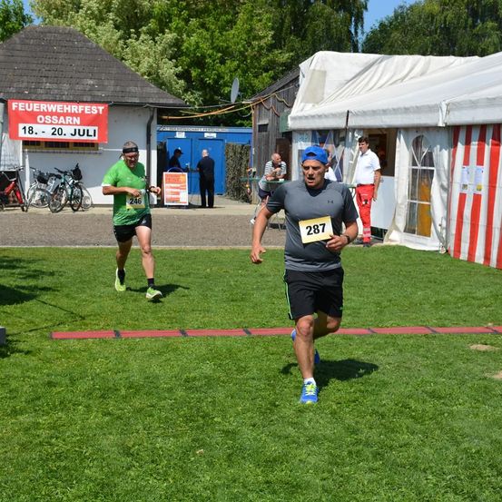 Zwei Läufer nehmen an einem Marathon auf einem Rasenfeld teil. Der Läufer vorne hat die Nummer 287 auf seinem Trikot, während der Läufer dahinter die Nummer 246 hat. Fahrräder sind auf der Seite geparkt, und ein Banner mit dem Text 'Feuerwehrfest Ossarn' hängt an einem Gebäude.
