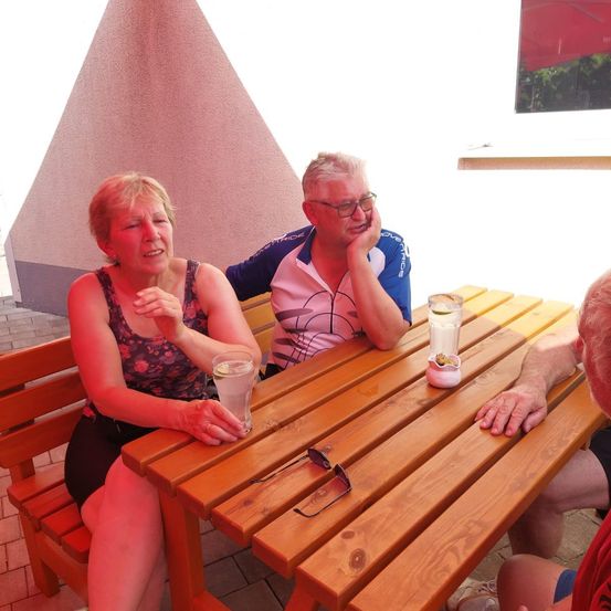 Vier Personen sitzen um einen Holztisch. Eine Frau lächelt und hält ein Getränk. Ein Mann stützt sein Kinn auf seine Hand. Ein anderer Mann stützt seinen Arm auf den Tisch. Eine Brille liegt auf dem Tisch.