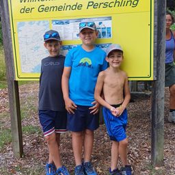 Drei Jungen stehen vor einem gelben Schild in einem Park und lächeln für die Kamera. Sie tragen Hüte und Turnschuhe. Eine Frau steht rechts, trägt eine Brille und ein ärmelloses Oberteil.