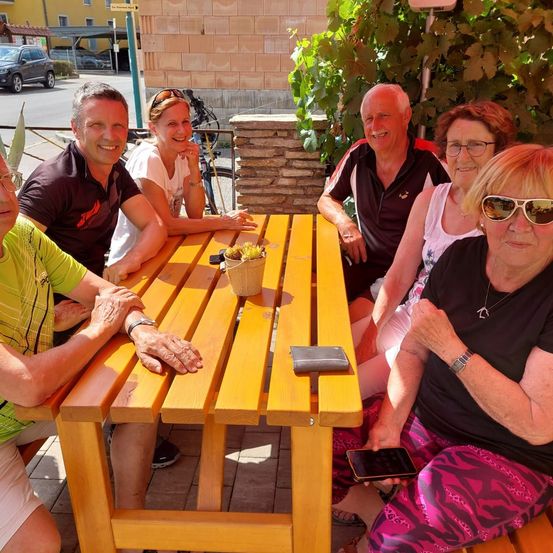 Eine Gruppe von Erwachsenen, sowohl Männer als auch Frauen, sitzen um einen Holztisch im Freien, lächeln und posieren für ein Foto. Auf dem Tisch steht eine Pflanze in einem Topf und ein Handy. Hinter ihnen steht eine Ziegelmauer mit Pflanzen.