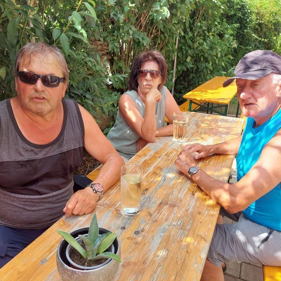 Drei Personen sitzen an einem Holztisch im Freien, möglicherweise in einem Garten. Ein Mann mit Sonnenbrille und ärmellosem Shirt sitzt links, eine Frau mit ärmellosem Shirt sitzt in der Mitte. Ein Mann in einem blauen Shirt und Hut sitzt rechts.