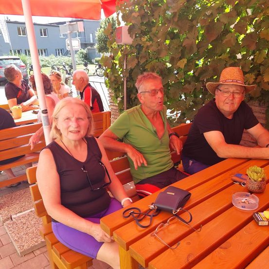 Drei Erwachsene sitzen unter einem roten Schirm an einem Außentisch, mit einer Frau im schwarzen Tanktop, einem Mann im grünen Shirt und einem Mann mit braunem Hut.