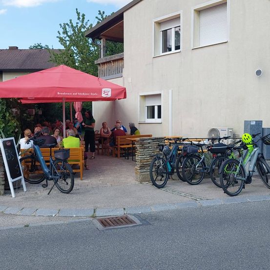 Eine Gruppe von Menschen sitzt unter einem roten Schirm um einen Tisch herum vor einem Gebäude. Fahrräder sind vor dem Gebäude geparkt.