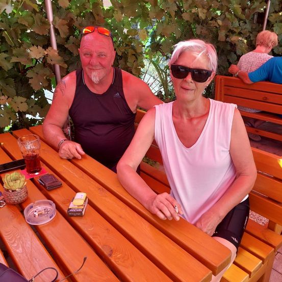 Ein älteres Paar sitzt auf einer Bank in einem Außenrestaurant. Der Mann trägt eine Sonnenbrille und die Frau raucht eine Zigarette. Der Tisch hat ein Glas mit Getränk, eine Brieftasche und eine Aschenschale. Dahinter ist ein verschwommener Blick auf eine andere Person, die auf einer Bank sitzt.