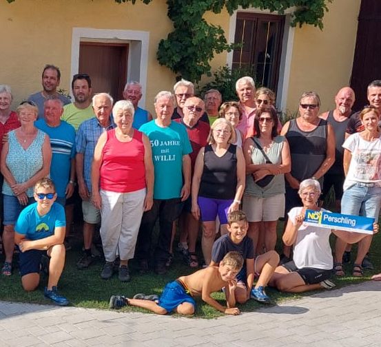 Eine große Gruppe von Erwachsenen und Kindern posiert für ein Foto vor einem gelben Gebäude mit Grünpflanzen. Einige halten ein Schild mit der Aufschrift NEIN! Perschling.