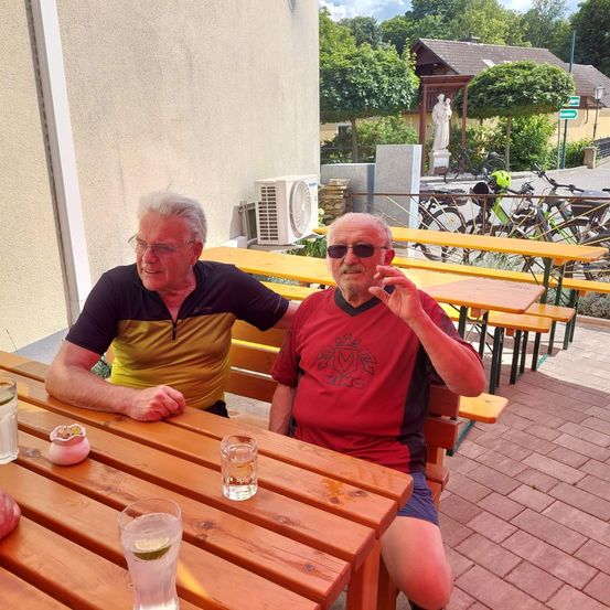 Zwei ältere Männer sitzen an einem Tisch im Freien und lächeln. Hinter ihnen sind Fahrräder geparkt und eine Statue steht. Auf dem Tisch befinden sich Gläser und ein rosa Kerzenhalter.