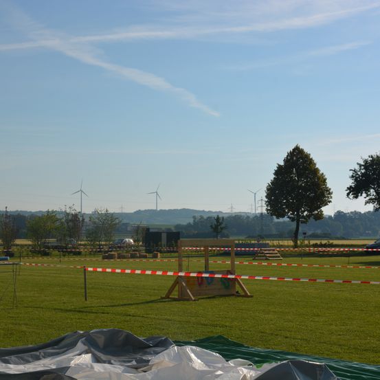 Ein Rasenfeld mit einer Holzstruktur, umgeben von Bäumen und Windrädern, hat eine rot-weiße Barriere. Der Himmel ist klar mit ein paar Wolken.
