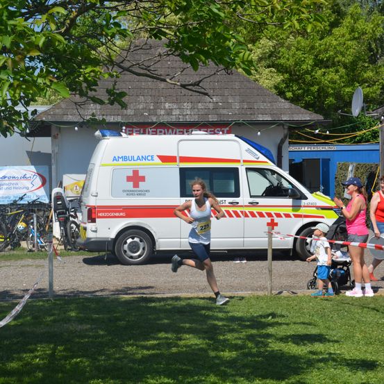 Eine Marathonläuferin beendet ihren Lauf in der Nähe eines Krankenwagens. Eine Frau und ein Kind stehen hinter einem Absperrband. Dahinter sind ein Gebäude und Fahrräder zu sehen.