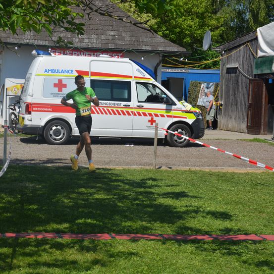 Ein Marathonläufer in Grün läuft vor einem Krankenwagen. Der Krankenwagen ist in der Nähe eines Gebäudes geparkt. Hinter dem Krankenwagen steht eine Person in der Nähe der Tür.