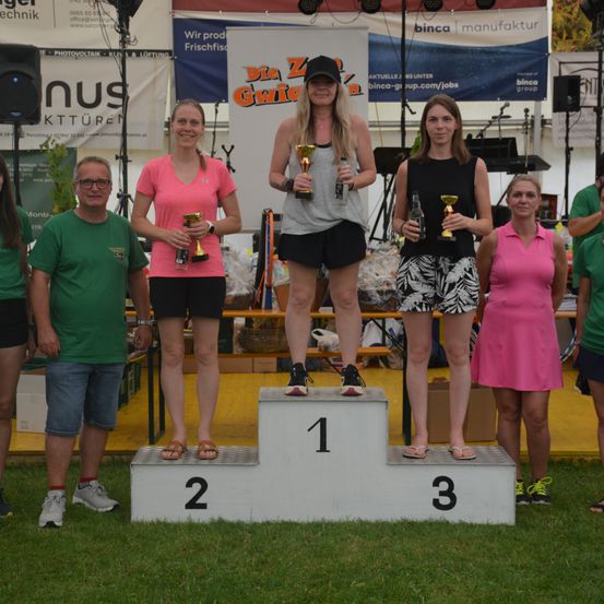Eine Gruppe von Menschen steht mit Trophäen auf einem Podest, zwei Frauen in der Mitte halten Trophäen und zwei Männer stehen links. Dahinter steht ein weißes Zelt.