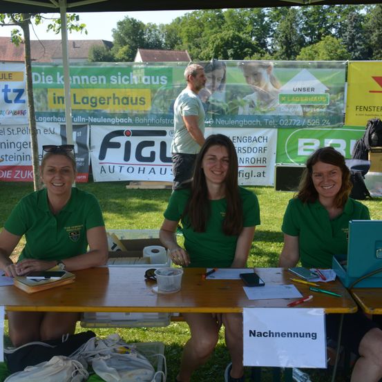 Drei Frauen in grünen Shirts sitzen an einem Tisch mit einem Schild, das 'Nachmeldung' steht. Sie lächeln und scheinen bei einer Veranstaltung zu arbeiten. Ein Mann steht hinter ihnen in der Nähe eines Schildes.