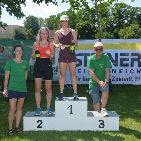 Vier Personen stehen auf einem Podium, zwei Frauen halten Flaschen, eine Frau hält eine Medaille und ein Mann sieht zur Seite. Hinter ihnen ist ein Banner.