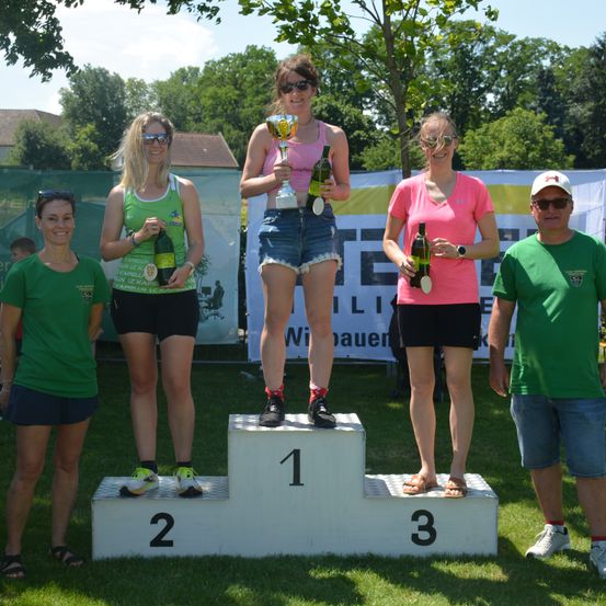 Fünf Frauen stehen auf einem Podium und halten Flaschen Wein und Trophäen. Sie tragen lässige Sportbekleidung, was auf eine Sportveranstaltung hindeutet. Hinter ihnen befindet sich ein Banner mit gelbem und weißem Text.