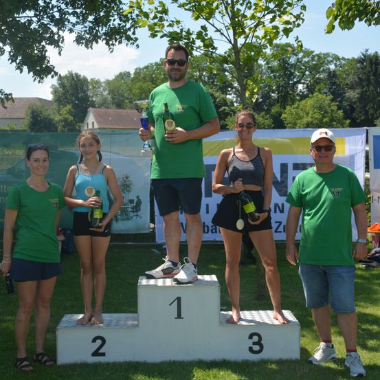 Ein Mann und drei Frauen stehen auf einem Podium mit den Nummern 1, 2 und 3. Sie tragen grüne Shirts und halten Flaschen und Pokale. Sie stehen auf einem Rasenfeld. Dahinter befindet sich ein Banner mit dem Wort 'EN'. In der Ferne sind Bäume und ein Gebäude zu sehen.