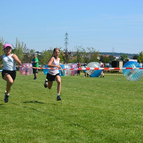Zwei Mädchen rennen auf einem Feld mit einer Ball-Hindernisparcours, andere beobachten im Hintergrund.
