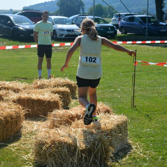 Eine Läuferin in einem weißen Shirt mit der Nummer 102 springt über Heuballen auf einem Grasfeld. Ein Mann in einem gelben Shirt steht hinter einem Hindernis.