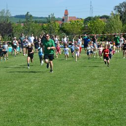 Eine Gruppe von Kindern und Erwachsenen rennt in einem Park. Einige tragen Hüte. Bäume und Gebäude sind im Hintergrund.