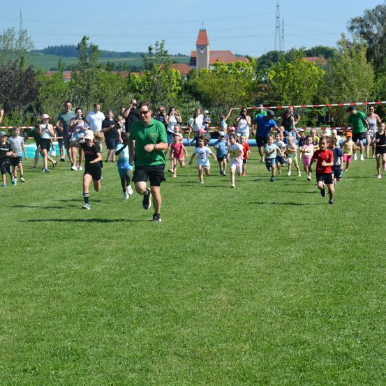 Eine Gruppe von Kindern und Erwachsenen rennt in einem Park. Einige tragen Hüte. Bäume und Gebäude sind im Hintergrund.