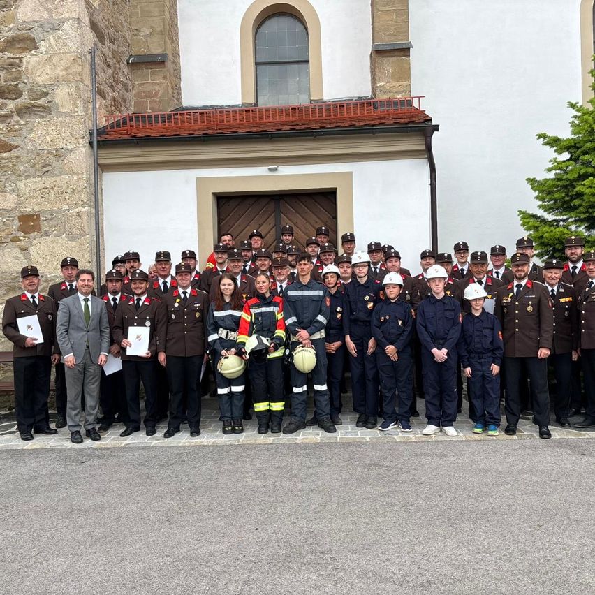 Eine Gruppe uniformierter Personen, darunter Feuerwehrleute und Offiziere, steht vor einem Gebäude aus Stein mit einem roten Dach und einem Baum in der Nähe.