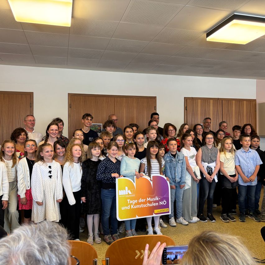 Eine Gruppe von Kindern und Erwachsenen steht in einem Raum und hält ein Schild mit der Aufschrift 'Tage der Musik- und Kunstschulen NO'. Sie tragen verschiedene Kleidungsstücke und einige haben Brillen. Der Raum hat Holztüren und Lichter an der Decke.