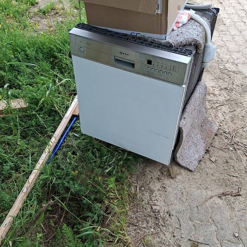 Ein Geschirrspüler steht auf dem Boden, bedeckt von einer braunen Box und einem Handtuch. Der Geschirrspüler hat ein Bedienfeld mit Tasten und einem silbernen Griff. Ein blauer Schlauch ist angeschlossen.