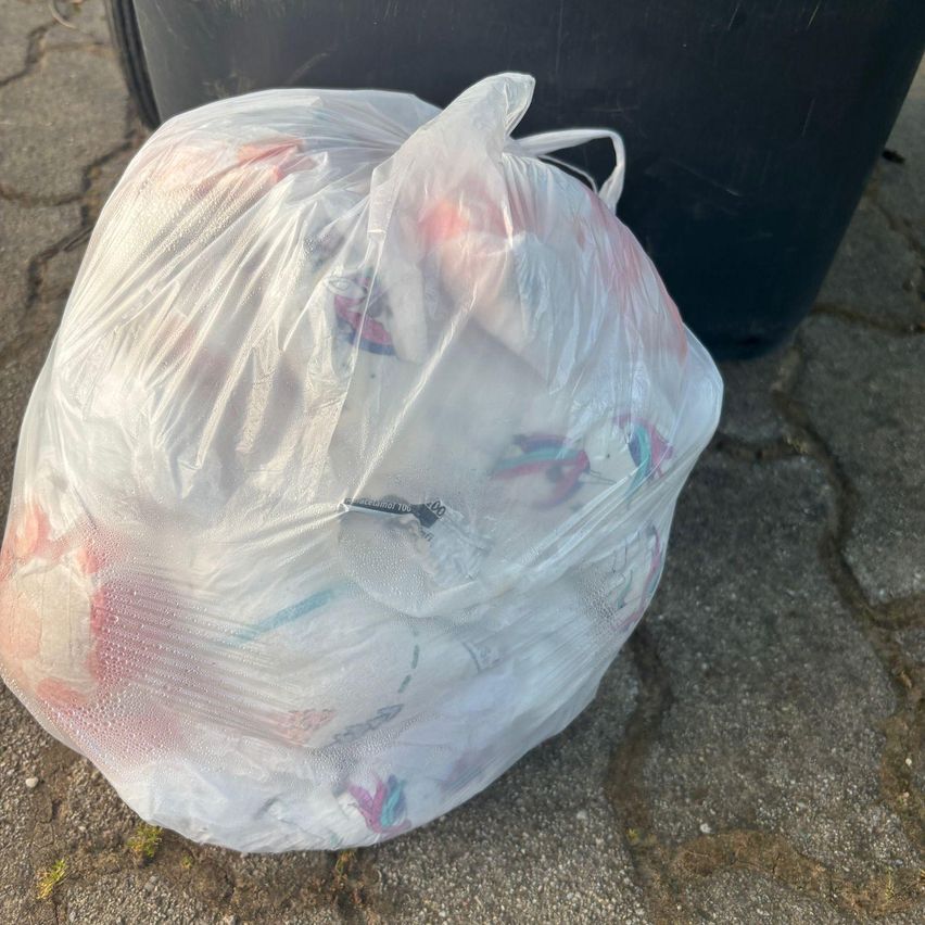 Ein durchsichtiger Plastiksack, gefüllt mit verschiedenen Gegenständen, liegt auf dem Boden neben einem schwarzen Mülleimer. Der sack ist aufgetrieben und enthält Artikel wie Kleidung, Lebensmittelverpackungen und möglicherweise einige persönliche Gegenstände.