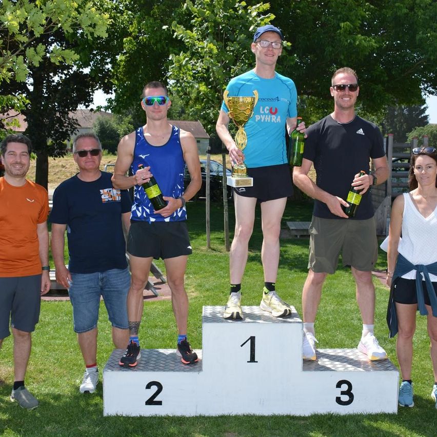 Eine Gruppe von Menschen auf einem Podium mit Flaschen, darunter ein Mann, der einen Pokal hält. Hinter ihnen hält ein Mann eine Flasche, während andere in Freizeitkleidung stehen. Sie befinden sich auf einem Rasenfeld mit Bäumen und einem Gebäude im Hintergrund.