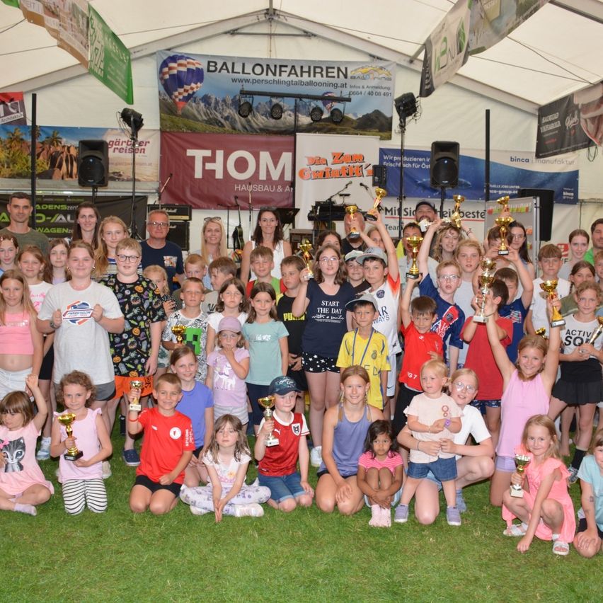 Eine Gruppe von Kindern und Erwachsenen posiert für ein Foto und hält Pokale. Sie befinden sich in einem Innenraum mit weißer Decke und Bannern. Einige von ihnen tragen Brillen und einige tragen Turnschuhe. Hinter ihnen steht ein Lautsprecher, und eine Person steht rechts.