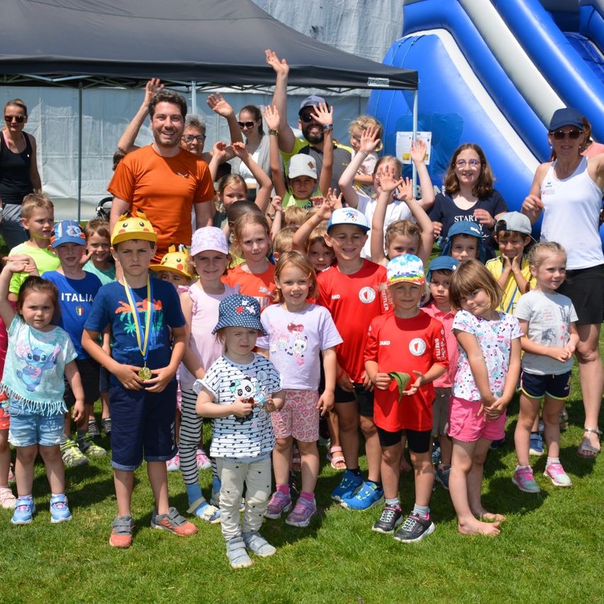 Eine Gruppe von Kindern und Erwachsenen posiert für ein Foto auf einem Rasenfeld mit einem blauen aufblasbaren Spielgerät und einem Zelt im Hintergrund. Einige tragen Medaillen und Hüte.