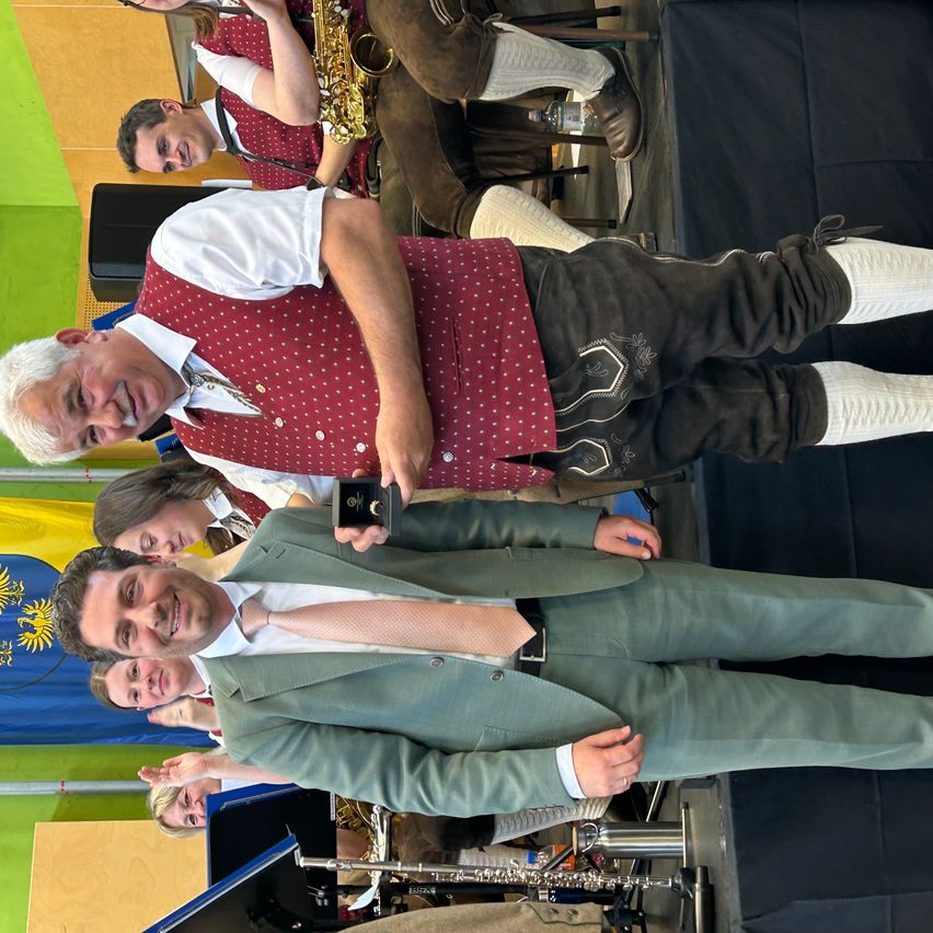 Eine Gruppe von Musikern, einer hält einen Pokal, auf einer Bühne mit einem farbenfrohen Hintergrund. Einige sind in traditioneller Kleidung gekleidet.