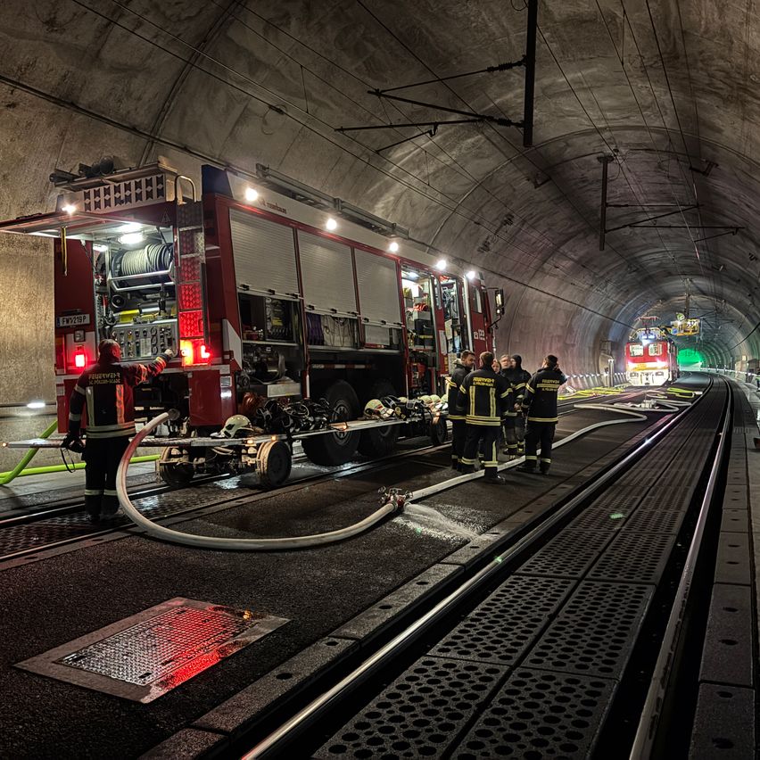 Ein Feuerwehrauto steht in einem Tunnel, mit Feuerwehrleuten in der Nähe. Ausrüstung und Schläuche sind ausgebreitet, und ein weiterer Zug ist am Ende des Tunnels sichtbar.