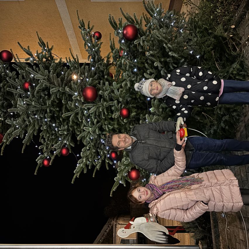Eine dreiköpfige Familie steht unter einem großen geschmückten Weihnachtsbaum, lächelt und posiert für ein Foto. Der Baum ist mit roten Kugeln und Lichtern geschmückt.