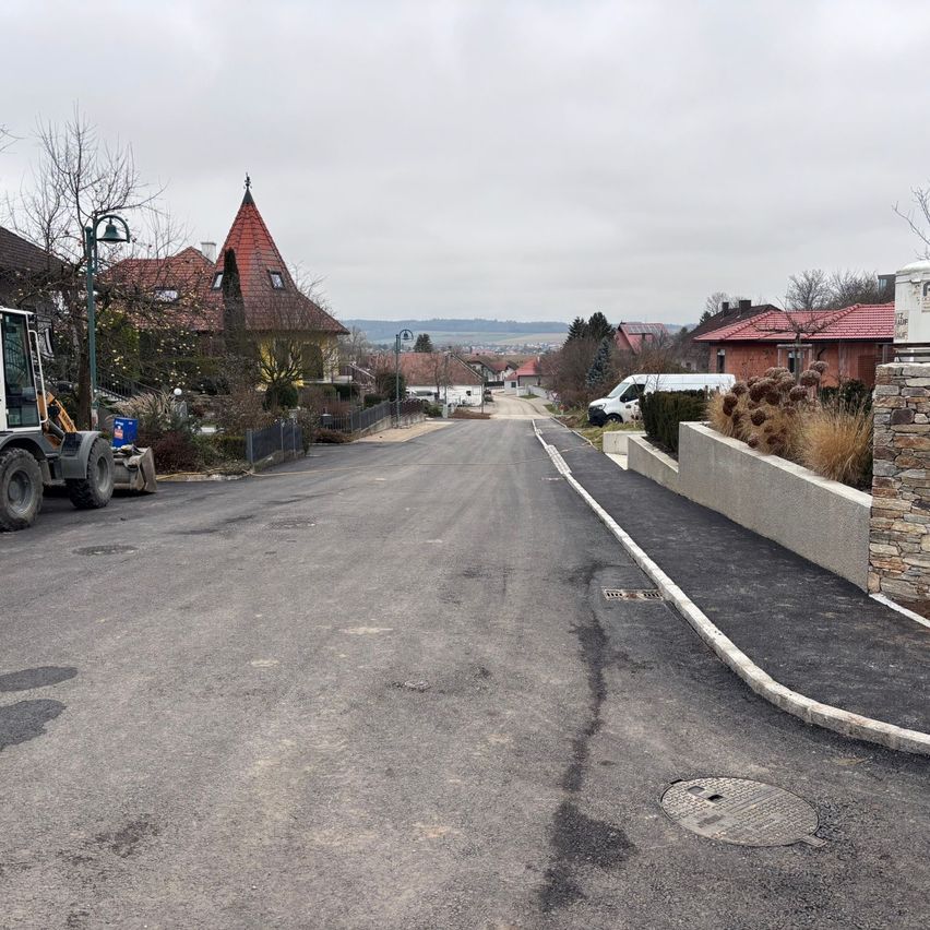 Eine leere Straße mit Häusern auf beiden Seiten, einem Baufahrzeug auf der linken Seite und einem geparkten weißen Lieferwagen. Bäume und Pflanzen sind vorhanden, und Berge sind in der Ferne sichtbar.