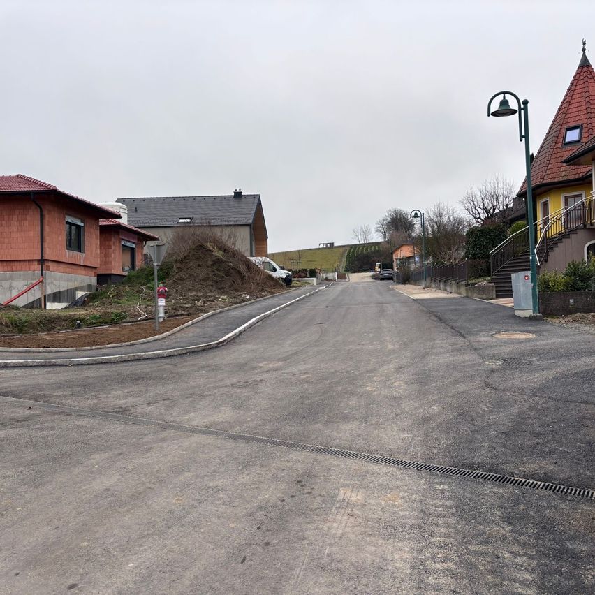 Eine leere Straße mit einer neuen Asphaltdecke, gesäumt von Häusern mit roten Dächern. Eine Baustelle ist sichtbar, mit einem Haufen Erde und einem geparkten weißen Lieferwagen. Straßenlaternen und ein Haus mit Treppe und Balkon befinden sich auf der rechten Seite.