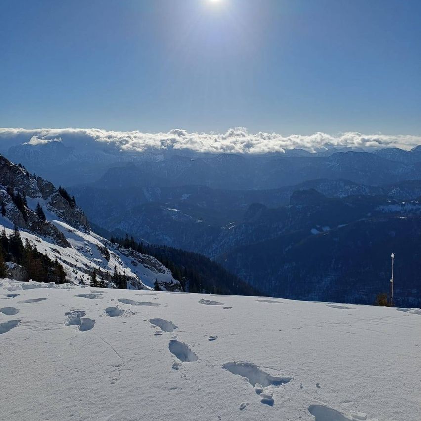 Ein schneebedeckter Berggipfel unter einem strahlenden Sonnenschein, mit Blick auf entfernte Berge und einen klaren Himmel. Fußabdrücke sind im Schnee sichtbar.