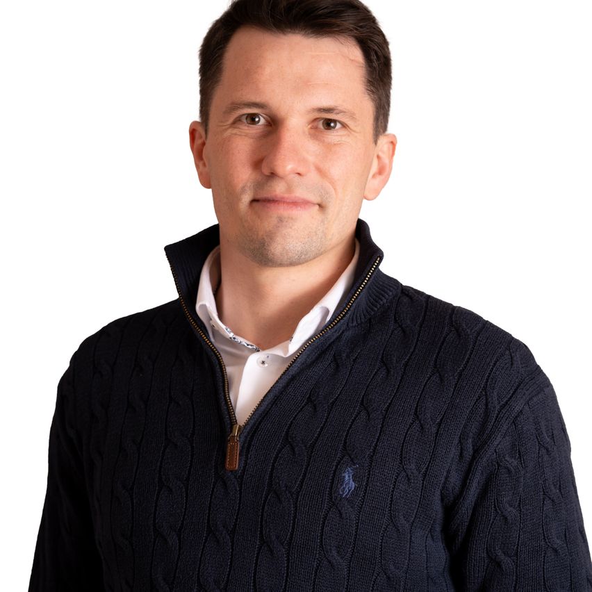 Ein Mann in einem dunkelblauen Pullover mit Logo auf der linken Seite und weißem Hemd darunter, vor weißem Hintergrund stehend.