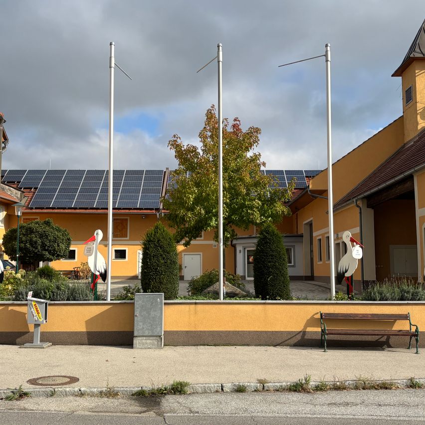 Ein Gebäude mit Solarmodulen auf dem Dach, umgeben von Grünflächen, zwei Storchskulpturen und einer Bank.