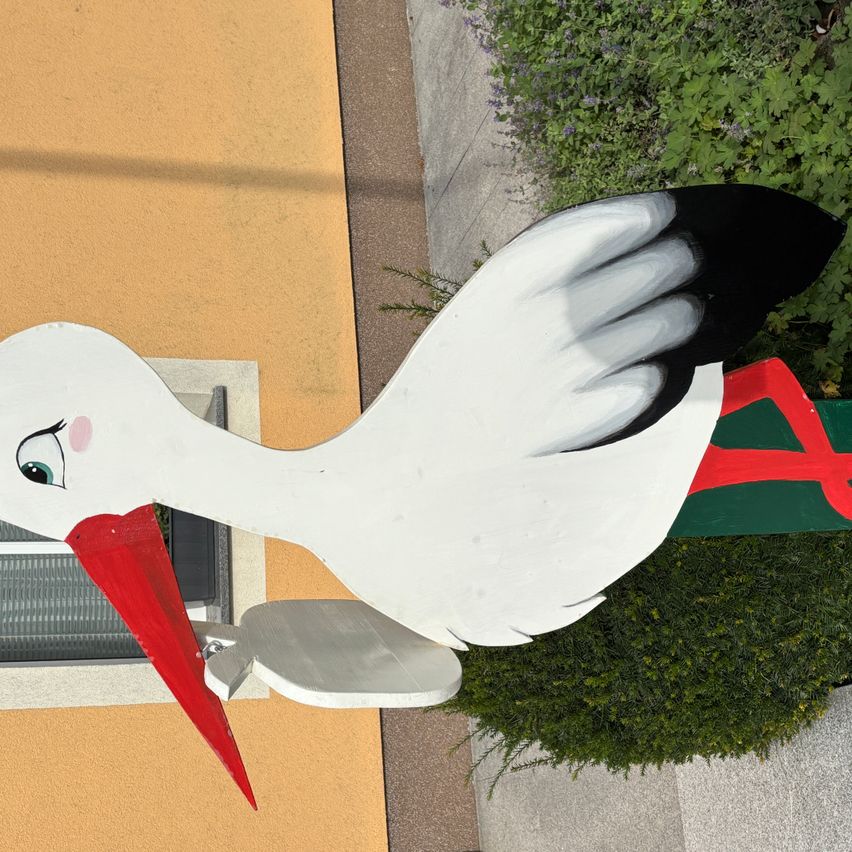 Ein hölzerner Storch mit rotem Schnabel und schwarz-weiß bemalten Flügeln ist an einem grünen und roten Schild an der Seite eines Gebäudes befestigt.